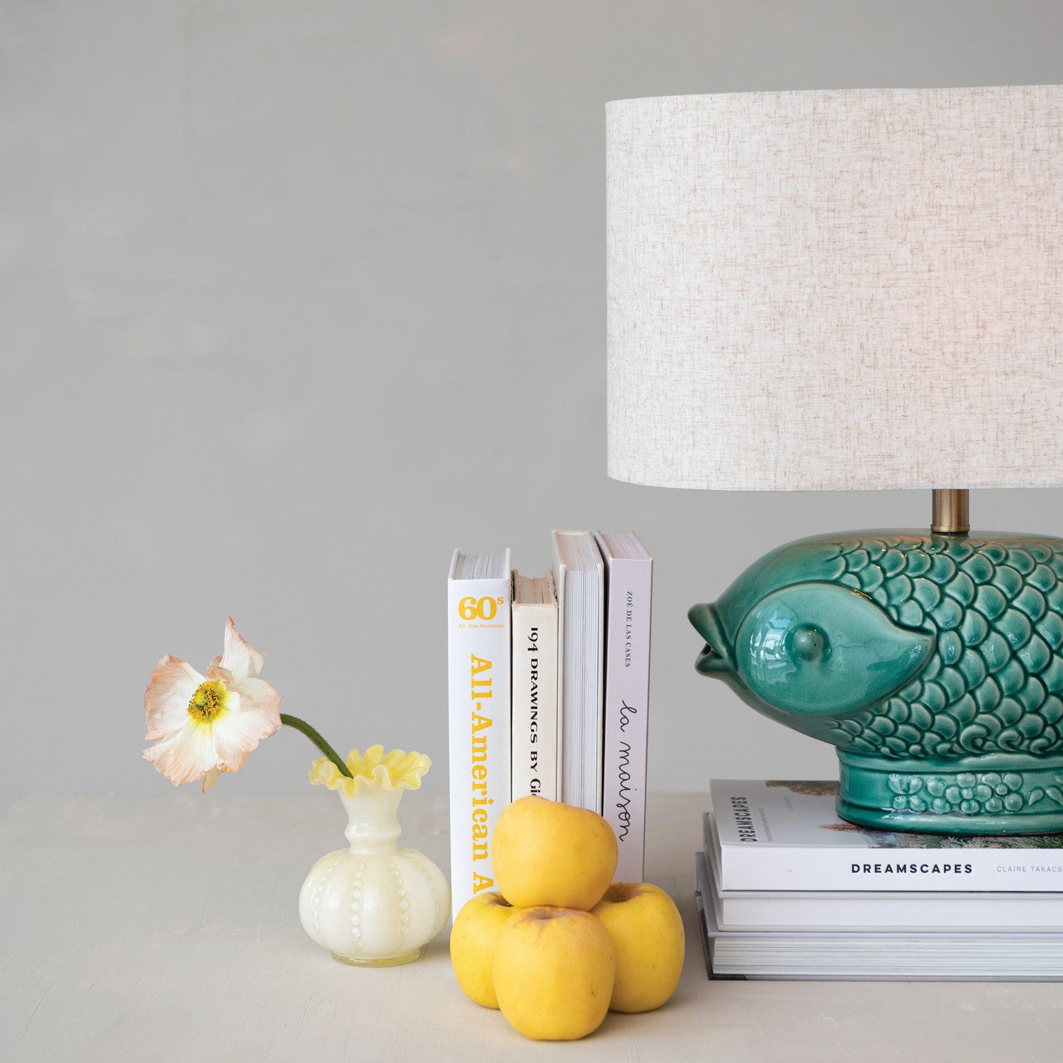 A styled interior scene with a teal ceramic fish-shaped table lamp beside a stack of books, a small white vase with a pale flower, and yellow fruit on a light surface against a soft gray background.