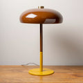 Modern table lamp with a glossy brown dome shade and yellow base on a wooden surface against a plain wall