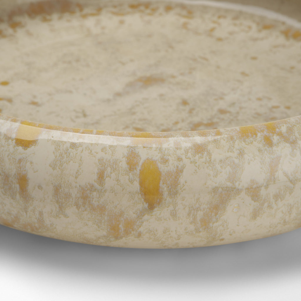 A close-up of a shallow round bowl or dish filled with a creamy off-white liquid or mixture, showing a marbled beige-and-cream surface with small yellowish inclusions.