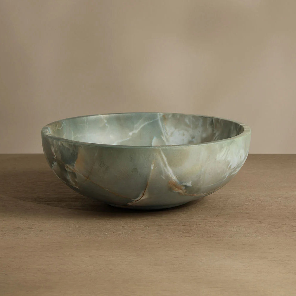 A round stone bowl with a greenish-gray marbled pattern and subtle gold veining, displayed on a wooden surface against a neutral beige background.