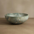 A round stone bowl with a greenish-gray marbled pattern and subtle gold veining, displayed on a wooden surface against a neutral beige background.