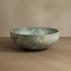 A round stone bowl with a greenish-gray marbled pattern and subtle gold veining, displayed on a wooden surface against a neutral beige background.