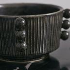 A close-up of a dark ceramic bowl with vertical ribbing and three glossy spherical handle details on the left side, set against a dark reflective surface.