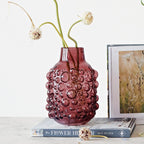 Purple glass vase with bubble texture holding dried flowers on books with floral art print nearby