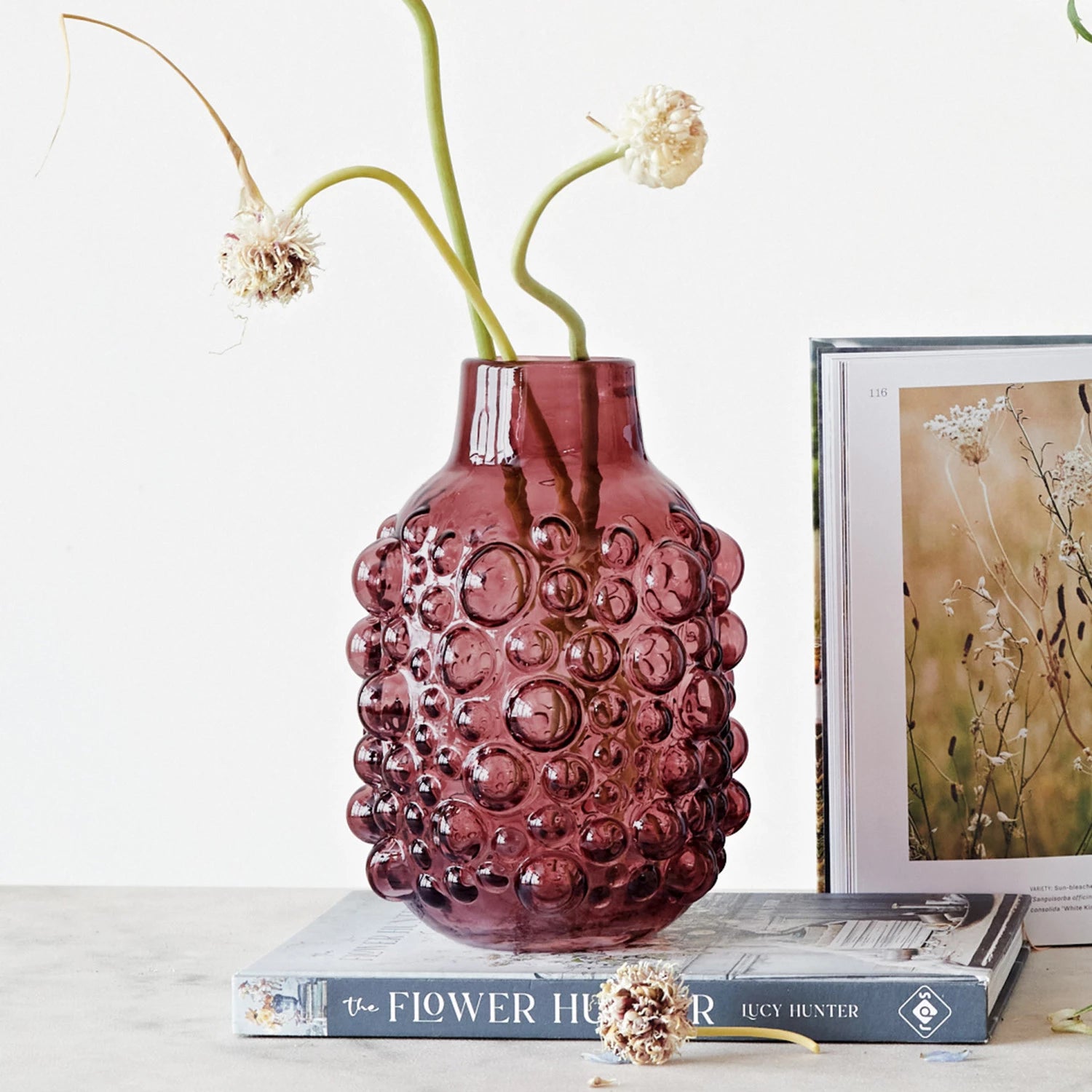 Purple glass vase with bubble texture holding dried flowers on books with floral art print nearby