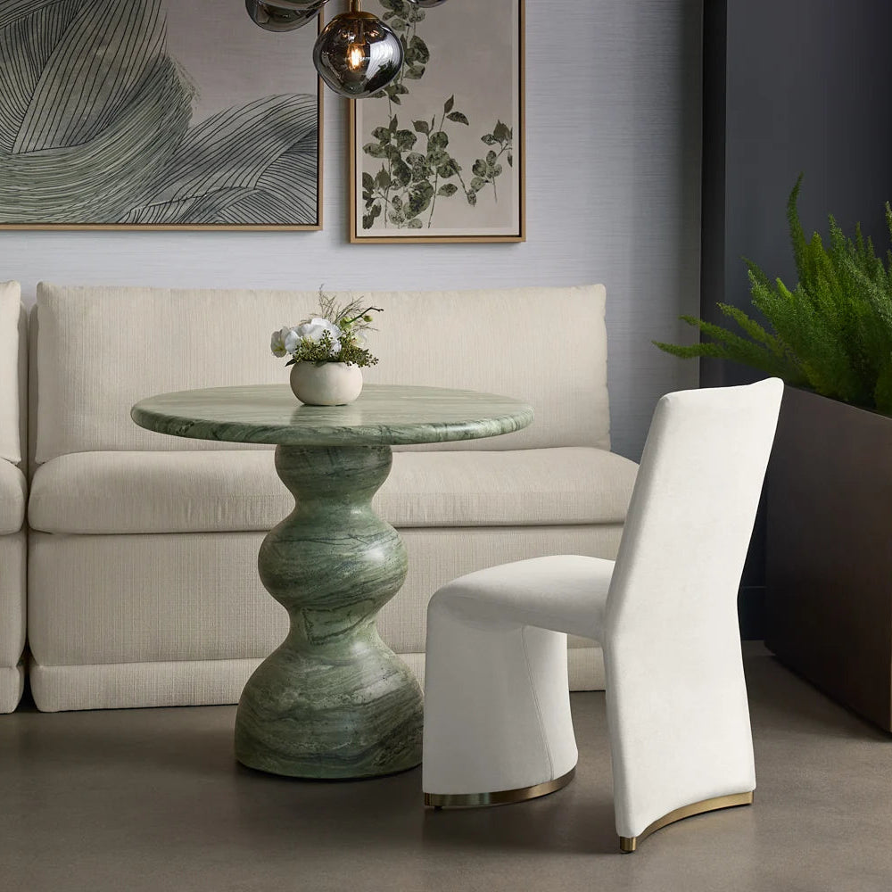 A modern living room scene featuring a round green marble pedestal table, a white upholstered accent chair, and a cream sectional sofa in the background.