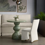 A modern living room scene featuring a round green marble pedestal table, a white upholstered accent chair, and a cream sectional sofa in the background.