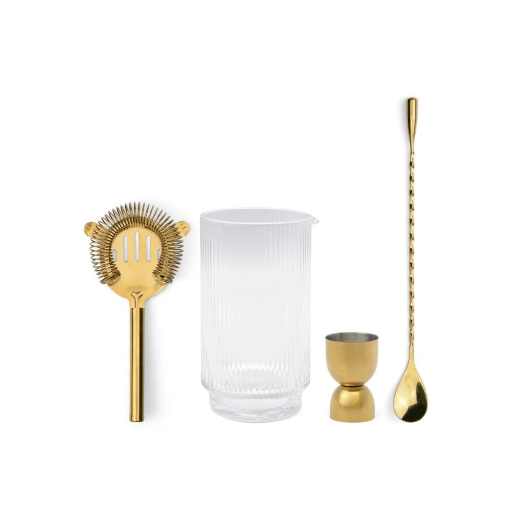 Set of gold cocktail tools including a strainer, jigger, and spoon with a clear glass on a white background.