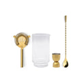 Set of gold cocktail tools including a strainer, jigger, and spoon with a clear glass on a white background.