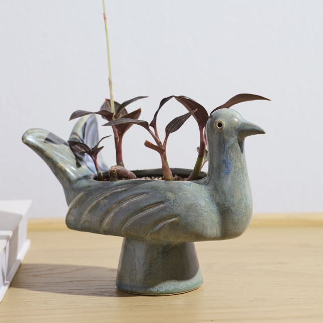 A ceramic bird-shaped planter in a muted blue-green glaze, holding several small dark-leafed plants, set on a light wood surface against a plain light gray wall.