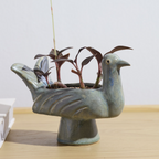A ceramic bird-shaped planter in a muted blue-green glaze, holding several small dark-leafed plants, set on a light wood surface against a plain light gray wall.