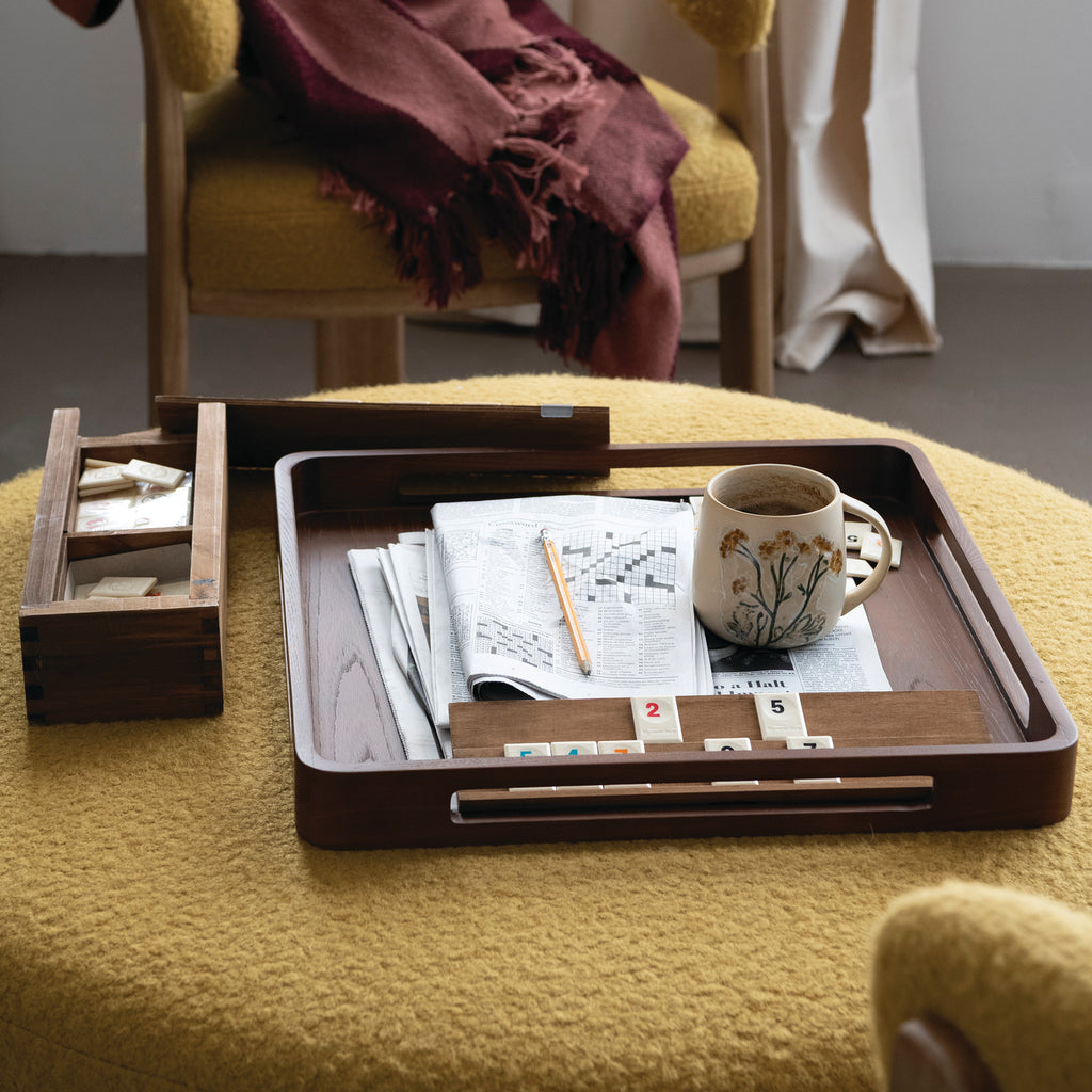 A dark brown wooden tray set on a yellow textured chair in a cozy room. The tray holds a folded newspaper, a white ceramic mug with a floral print, a pencil, and a small set of white tiles.