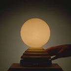A glowing spherical table lamp with a ribbed tiered base, shown on a dark surface against a dim background, with a hand reaching toward the right side of the lamp.