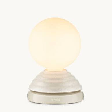 A modern table lamp with a glowing warm white spherical globe on a textured, tiered off-white base with a digital clock display showing 07:00.