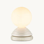 A modern table lamp with a glowing warm white spherical globe on a textured, tiered off-white base with a digital clock display showing 07:00.