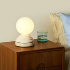A lit spherical table lamp with a ribbed cream-colored base sits on a wooden nightstand next to a glass of water, with a bed and pillows visible in the softly lit bedroom setting.