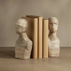 Two beige stone bust bookends holding a set of tan books on a wooden surface against a neutral beige background.