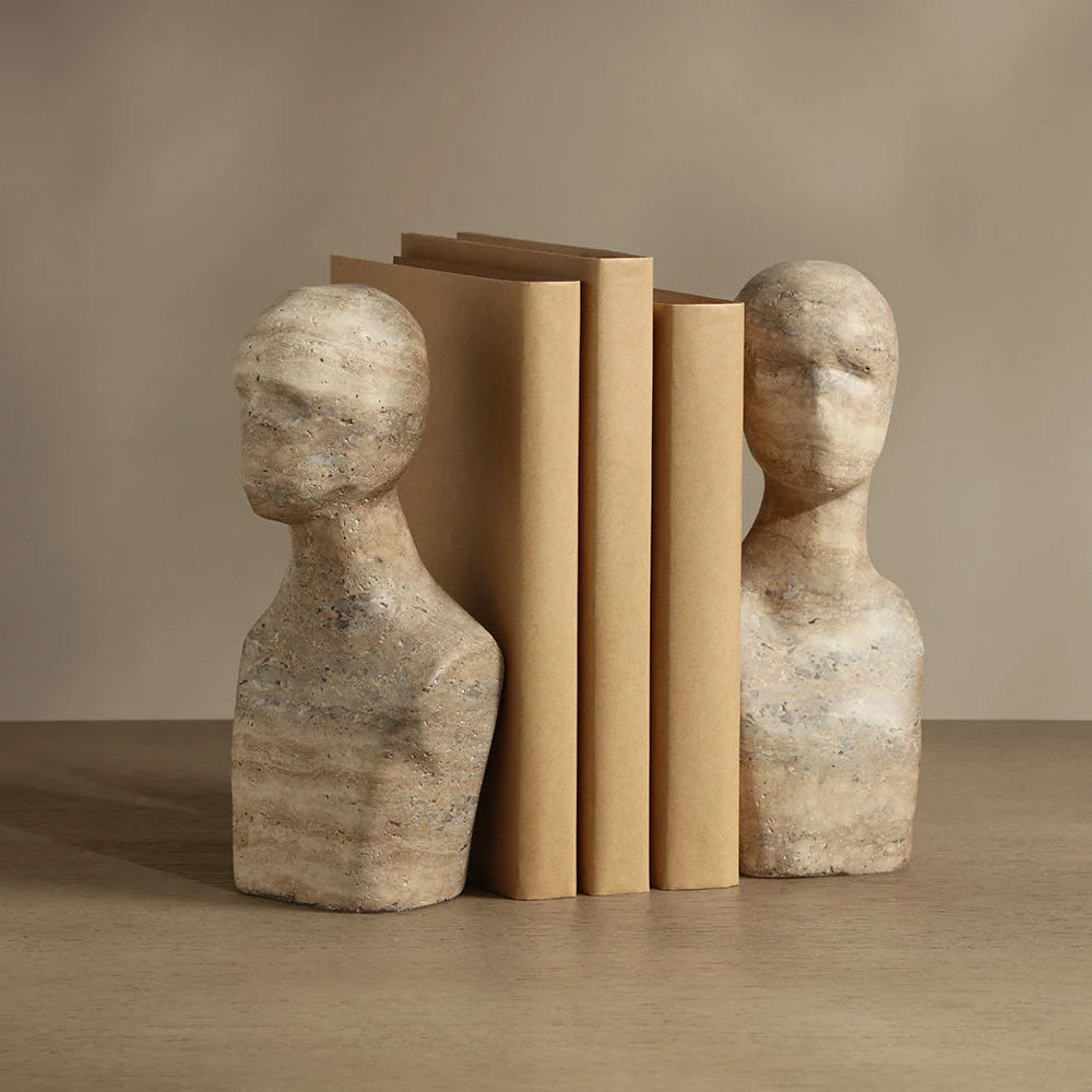 Two beige stone bust bookends holding a set of tan books on a wooden surface against a neutral beige background.